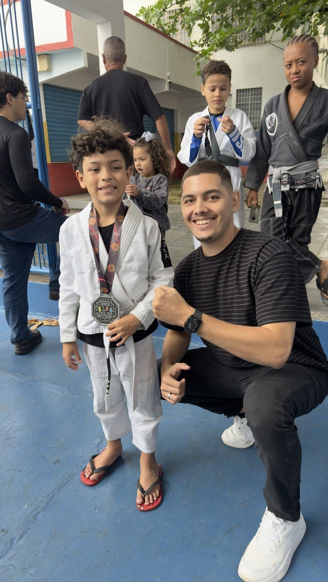 Thiago Antunes ao lado de uma criança de kimono branco com uma medalha de jiu-jitsu