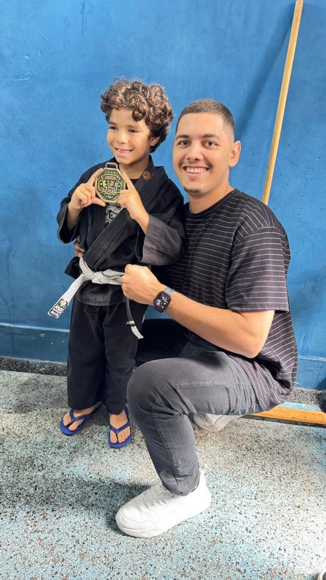 Thiago Antunes ao lado de uma criança de kimono preto com uma medalha de jiu-jitsu